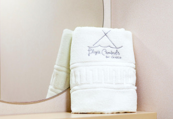 Toalla blanca doblada con bordado 'Playa Cambrils by Casillo' sobre una mesa junto a un espejo en el lodge.