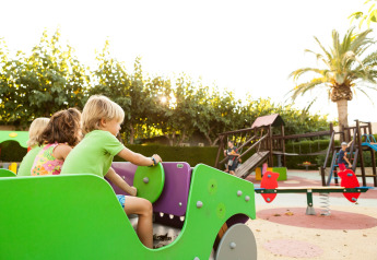 Kinderen spelen op een kleurrijke speeltuin bij een glampingaccommodatie, omringd door bomen en palmen.