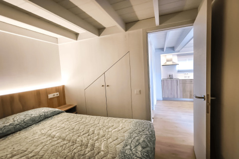 Soveværelse med synlige bjælker og seng, set mod et moderne køkken i La Llosa 1 Xaloc, Spanien.