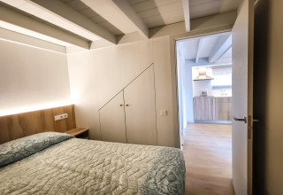 Soveværelse med synlige bjælker og seng, set mod et moderne køkken i La Llosa 1 Xaloc, Spanien.
