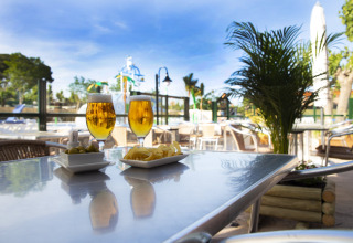 Deux verres de bière et des snacks sur une table extérieure dans un glamping avec vue sur piscine et verdure.