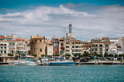 Vue sur un port pittoresque avec voiliers, tour en pierre et hébergement glamping moderne en bord de mer.