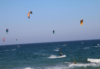 Scène de kitesurf colorée sur la mer près d’un glamping, cerfs-volants flottant sous un ciel bleu.