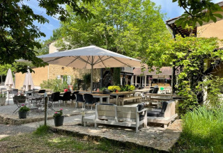 Zona de terraza exterior en Camping Le Clou 1, Francia, con mesas, sombrillas y vegetación frondosa.
