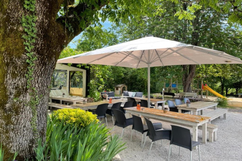 Zona exterior de Camping Le Clou 1 en Francia con sombrilla, mesas de madera, sillas y parque infantil al fondo.