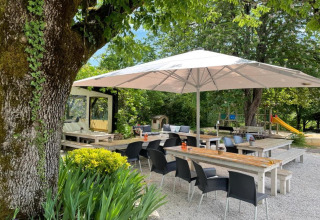 Zona exterior de Camping Le Clou 1 en Francia con sombrilla, mesas de madera, sillas y parque infantil al fondo.