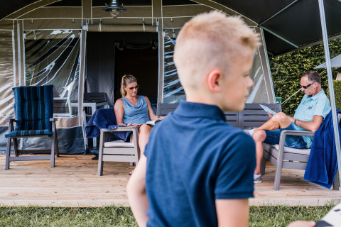 Familia relajándose en la terraza de una tienda safari, leyendo, con un niño en primer plano en verano.