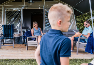 Familia relajándose en la terraza de una tienda safari, leyendo, con un niño en primer plano en verano.