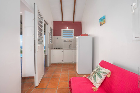 Acogedora cocina de glamping con nevera, estufa, sofá rojo y cojín tropical sobre suelo de terracota.