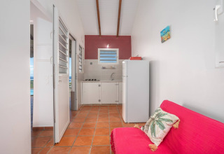 Acogedora cocina de glamping con nevera, estufa, sofá rojo y cojín tropical sobre suelo de terracota.