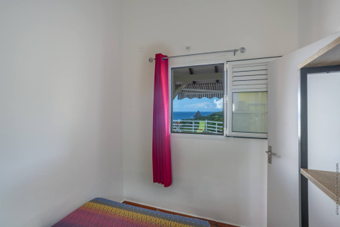 Habitación con colcha colorida, ventana abierta, cortina rosa y vista de montaña y mar en Portugal.