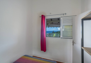 Habitación con colcha colorida, ventana abierta, cortina rosa y vista de montaña y mar en Portugal.