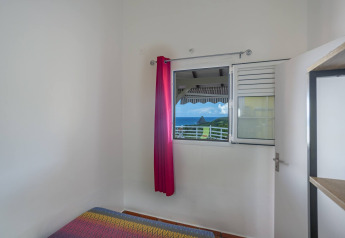Zimmer mit buntem Bettüberwurf, offenem Fenster, rosa Vorhang und Blick auf Berge und Meer in Portugal.