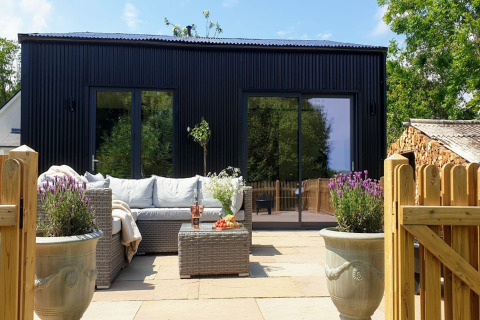 Moderno lodge nero a Blackbarn Wood con patio, divano in rattan, piante in vaso e cancello di legno.