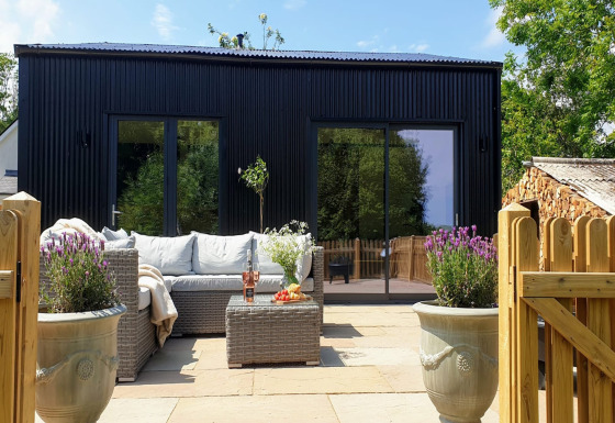 Moderno lodge nero a Blackbarn Wood con patio, divano in rattan, piante in vaso e cancello di legno.