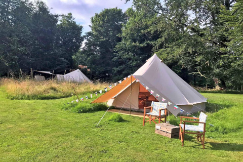 A large teepee-style tent on grass, with two chairs and a small box, surrounded by green trees.