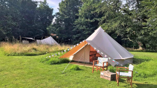 A large teepee-style tent on grass, with two chairs and a small box, surrounded by green trees.