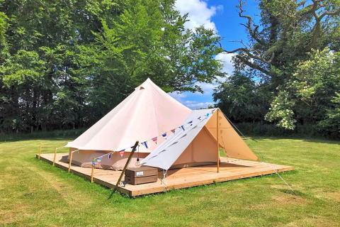 A teepee tent named Meadow Glamping Tents set up on a wooden platform in a grassy field with trees behind.