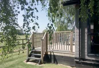 Vue ensoleillée sur la terrasse en bois à New Puddledock Lodge, Royaume-Uni, entourée de pelouse et d’arbres.
