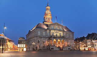 Beleuchtetes historisches Gebäude auf einem Platz in der Abenddämmerung, umgeben von moderner Architektur.