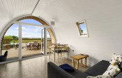 Lichte woonkamer in The Chicken Coop pod met gebogen wand, bank, eettafel en houten terras met uitzicht.