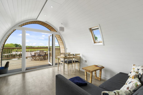 Lichte woonkamer in The Chicken Coop pod met gebogen wand, bank, eettafel en houten terras met uitzicht.