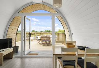 Interieur van een pod met eettafel en glazen deuren naar een houten terras met buitentafel en stoelen.