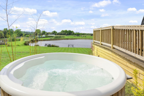 Glamping-accommodatie met een bubbelbad, houten terras en uitzicht op een meer en groene weide.