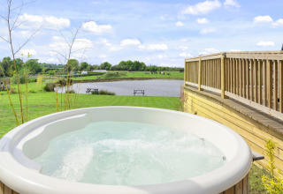 Glamping-accommodatie met een bubbelbad, houten terras en uitzicht op een meer en groene weide.