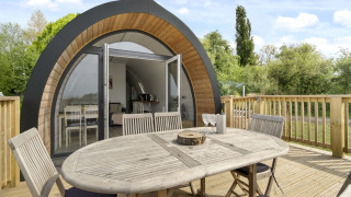 Houten tafel en stoelen op het terras voor de gebogen houten pod The Chicken Coop met glazen voordeur.