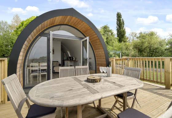 Houten tafel en stoelen op het terras voor de gebogen houten pod The Chicken Coop met glazen voordeur.