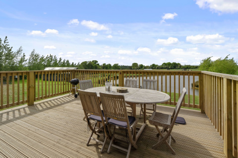 Houten terras bij een pod met buitentafel, stoelen, barbecue en uitzicht op een meer en weilanden.