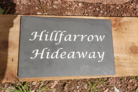 Et skilt med teksten 'Hillfarrow Hideaway' udenfor en hytte i Storbritannien, på en baggrund af træ og jord.