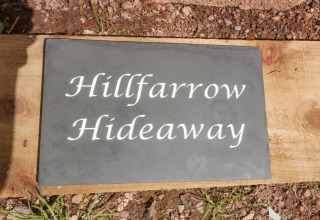 Et skilt med teksten 'Hillfarrow Hideaway' udenfor en hytte i Storbritannien, på en baggrund af træ og jord.