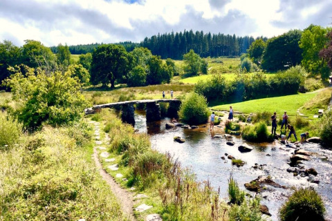 Billede af en malerisk glampingplads med stenbro over en strøm, grønne marker og mennesker i naturen.