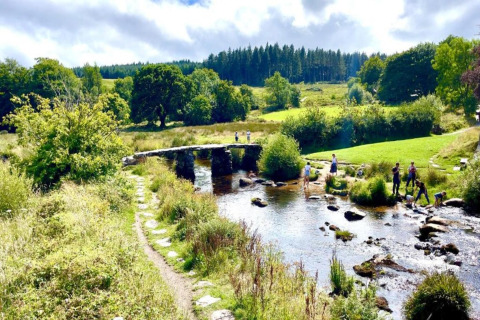Billede af en malerisk glampingplads med stenbro over en strøm, grønne marker og mennesker i naturen.