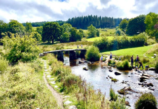 Billede af en malerisk glampingplads med stenbro over en strøm, grønne marker og mennesker i naturen.