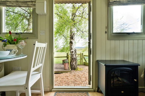 Interno di una cabina accogliente con porta aperta e vista sull’albero e campagna a Hay and Hedgerow Glamping UK