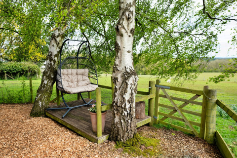 Area relax all'aperto a Hay and Hedgerow Glamping con sedia a dondolo, pedana in legno e prato.
