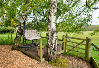 Area relax all'aperto a Hay and Hedgerow Glamping con sedia a dondolo, pedana in legno e prato.