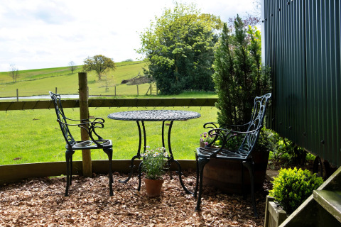 Tavolo e due sedie nere in metallo all’aperto con vista sui prati verdi di Hay and Hedgerow Glamping, Regno Unito.