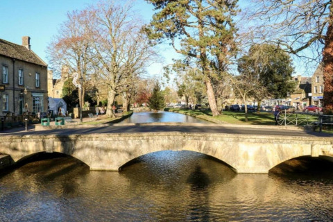 Stenen brug over een rustige rivier bij Little Rissington met bomen en gebouwen eromheen, Verenigd Koninkrijk.