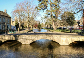 Stenen brug over een rustige rivier bij Little Rissington met bomen en gebouwen eromheen, Verenigd Koninkrijk.