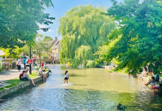 Mensen genieten aan de ondiepe rivier bij Little Rissington - Barn Four, omringd door groen en bomen.