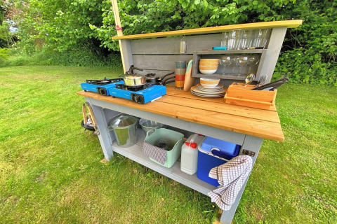 Outdoor cooking station by a teepee tent, with gas stoves, dishes, glasses, and a cooler on the grass.