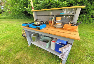 Outdoor cooking station by a teepee tent, with gas stoves, dishes, glasses, and a cooler on the grass.