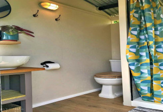 Modern bathroom inside a teepee tent with toilet, sink, mirror, wooden shelving and colorful shower curtain.