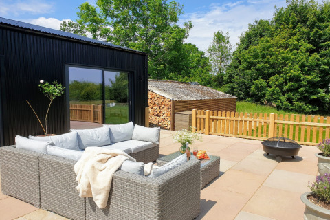 Zona salotto esterna al Blackbarn Wood lodge in UK con divano in rattan, braciere e vista verde.