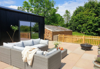 Zona salotto esterna al Blackbarn Wood lodge in UK con divano in rattan, braciere e vista verde.
