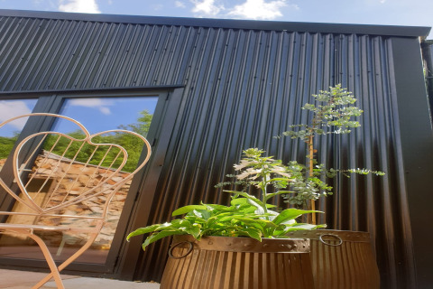 Capanna in legno nero con finestra riflettente, pianta in vaso e sedia in metallo a Blackbarn Wood, Regno Unito.